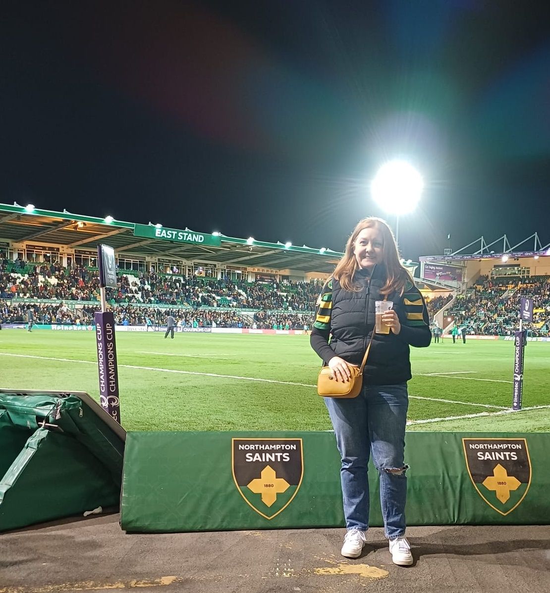 Sally standing pitchside at Franklin’s Gardens during an evening rugby match, holding a drink and wearing a Northampton Saints scarf. The rugby pitch and goalposts are behind them, with a packed stand lit by floodlights and “Northampton Saints” signage visible on the pitch barrier.