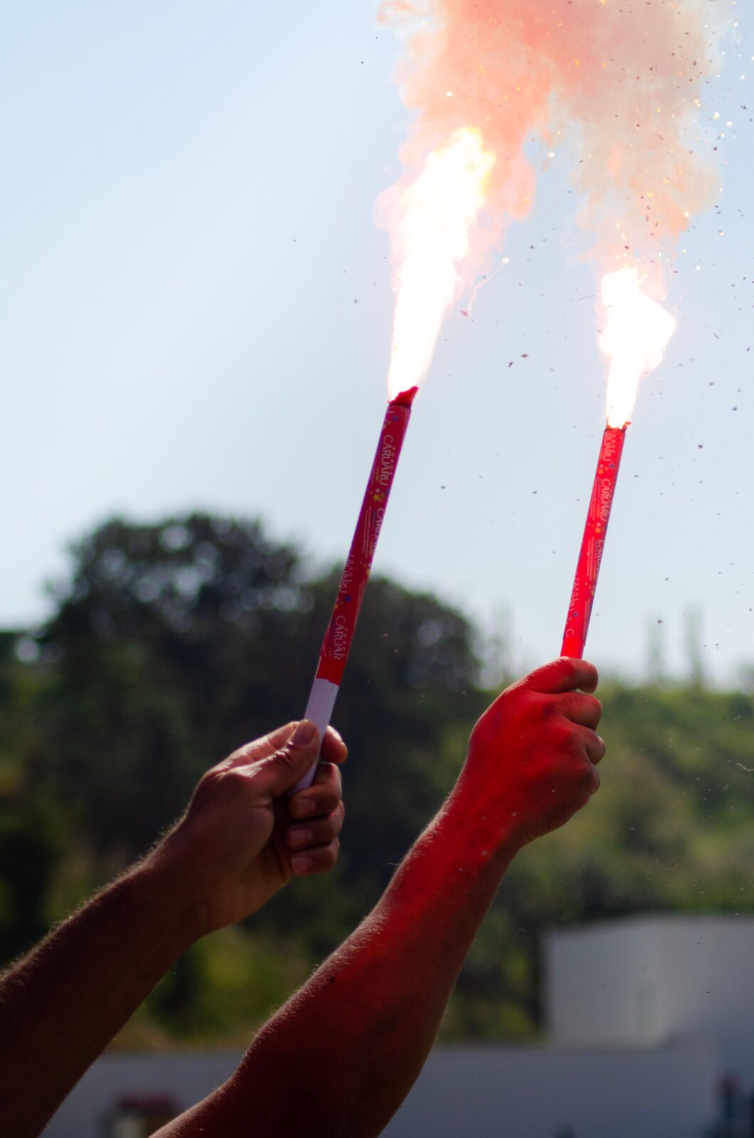 Two hands holding red flares with smoke on a sunny day outdoors, trees in the background.